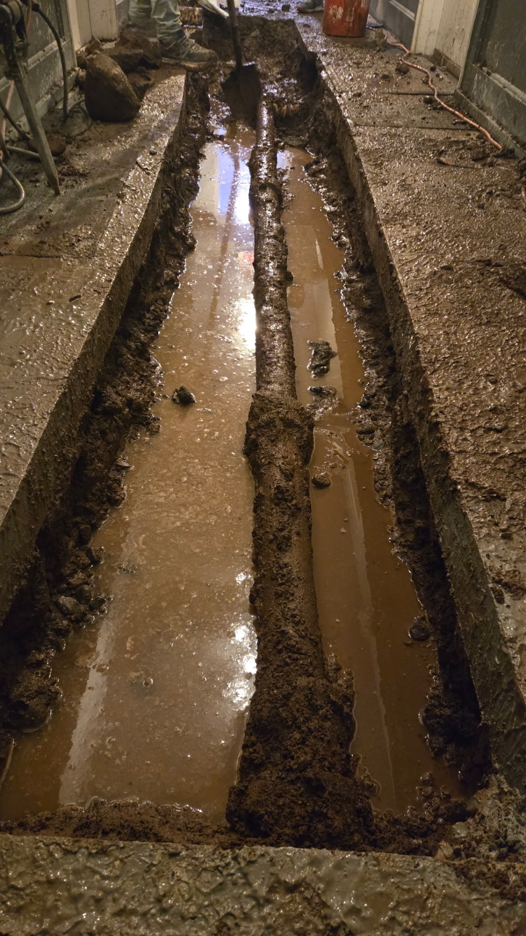 Excavation through the concrete slab to expose the full run of the failed drain line — carefully cut, shored, and dewatered inside an occupied building.