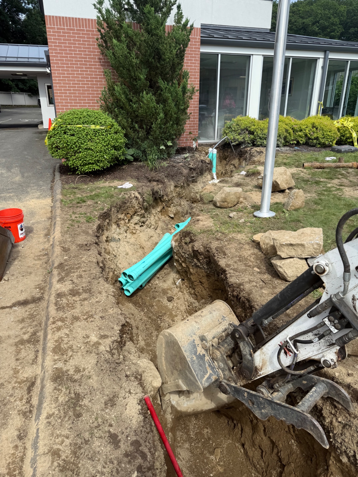 Trench opened on a commercial property front lawn — stockpiled new PVC on-site, mini-excavator working carefully around landscaping and the building foundation.