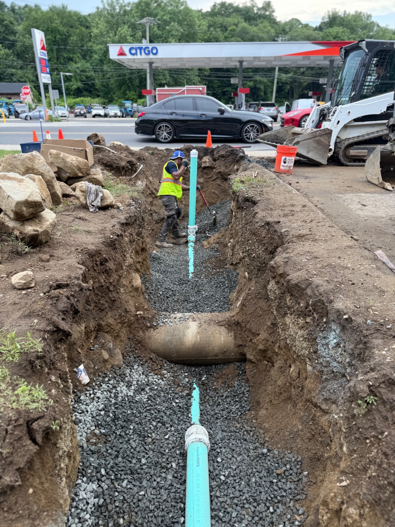 Commercial sewer replacement in progress along a busy CT roadway — our tech dressing the trench beside the Drain Wizard bucket while a skid-steer stands by. Traffic cones out, safety first.